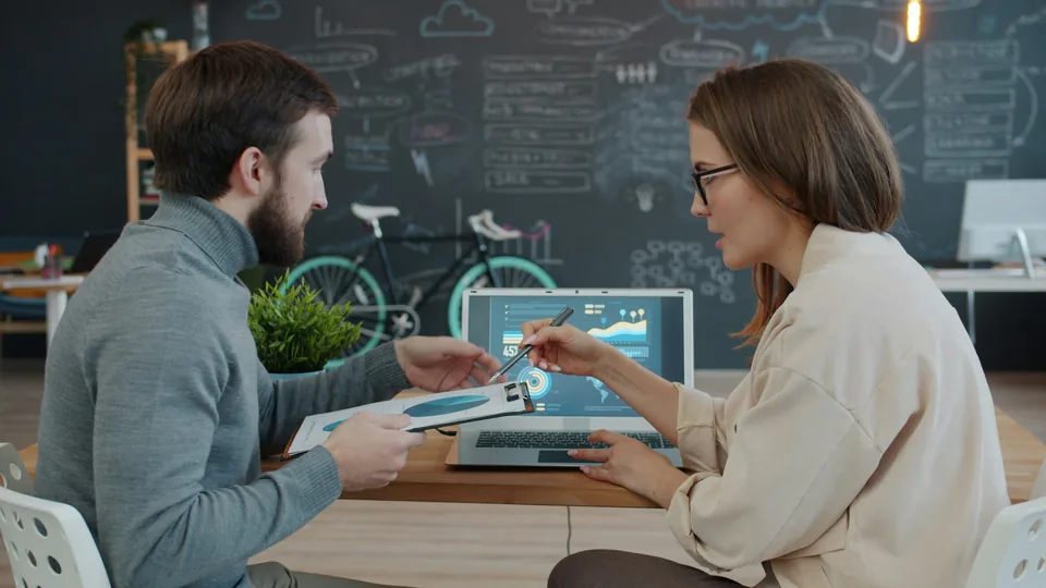 Colleagues review information together on a laptop in a business setting.